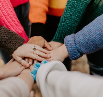 close up of 8 hands piled atop each other in a circle, each person wears a different colour jumper