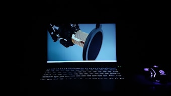 A dark setting featuring an illuminated laptop screen displaying a large, detailed image of a studio microphone with a pop filter. The keyboard is softly lit, glowing in the dark. To the right, a gaming mouse emits a faint purple light, adding to the overall moody atmosphere.