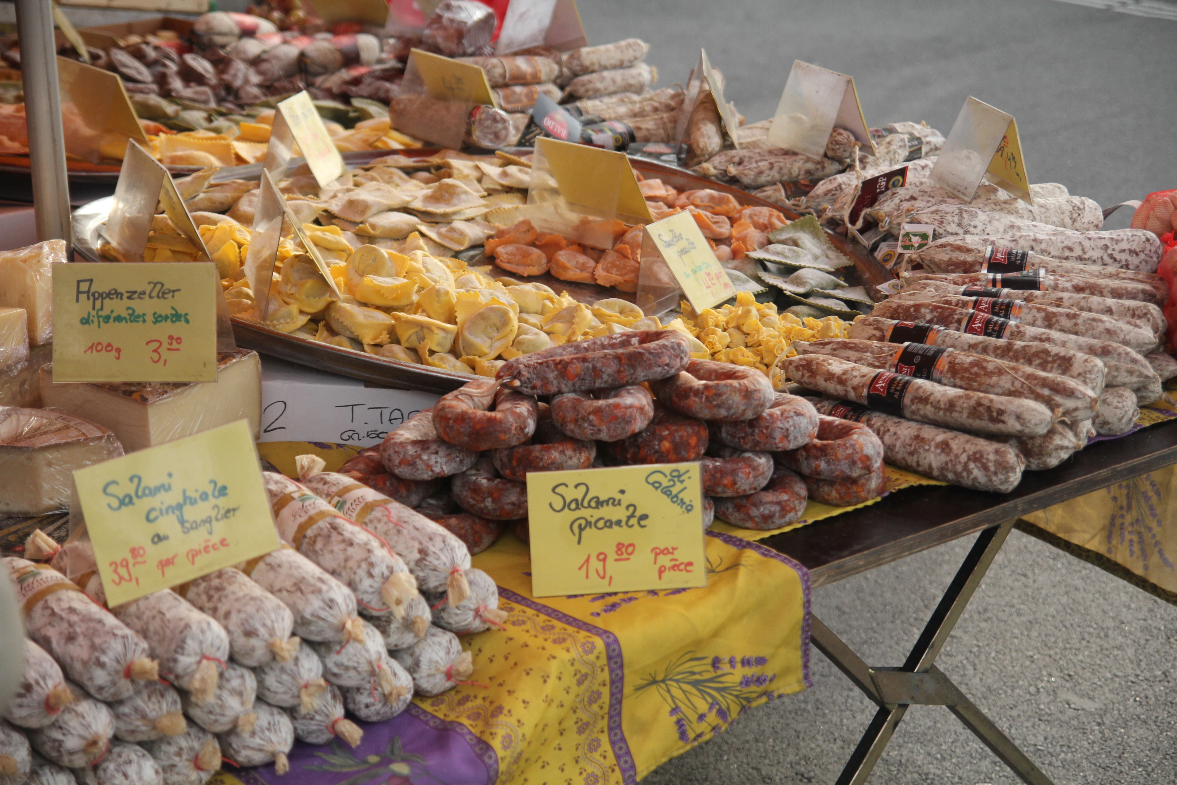 A charcuterie stall at a Geneva farmers' market, its table piled with cured salami and dried sausages of every size beside handwritten French price cards and slabs of cheese.
