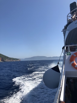 A boat navigates through clear blue waters with a visible wake trailing behind. On the right side of the image, part of the boat is visible with a life buoy and a large fender attached. The horizon reveals a distant view of green hills and mountainous islands under a cloudless sky.