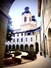 An elegant historical building with arched windows and a clock tower. The foreground features a manicured garden with neatly trimmed hedges and gravel paths. The architecture is classical, with stone facades and ornamental details.