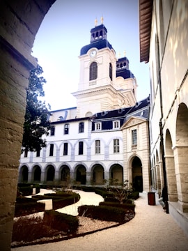 An elegant historical building with arched windows and a clock tower. The foreground features a manicured garden with neatly trimmed hedges and gravel paths. The architecture is classical, with stone facades and ornamental details.