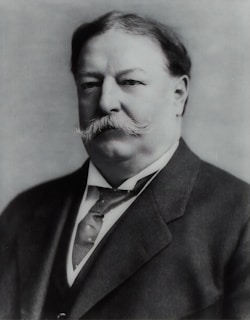 A black and white portrait of a distinguished man with a mustache, wearing a suit and tie. The image has a formal and historical feel.