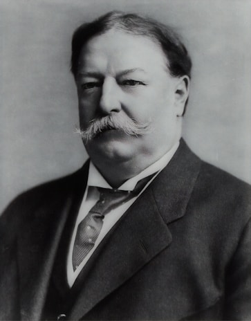 A black and white portrait of a distinguished man with a mustache, wearing a suit and tie. The image has a formal and historical feel.