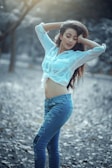 A woman stands in a natural outdoor setting with her arms raised and hands resting on her head. She is wearing a white blouse tied at the waist and distressed blue jeans. The background consists of trees and a ground covered with leaves, creating a serene atmosphere.