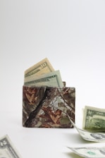 A camouflage-patterned wallet is open, with visible US dollar bills partially sticking out. Additional bills are scattered on the flat surface in front of the wallet, with a plain white background giving a minimal and clean appearance.