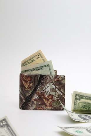 A camouflage-patterned wallet is open, with visible US dollar bills partially sticking out. Additional bills are scattered on the flat surface in front of the wallet, with a plain white background giving a minimal and clean appearance.