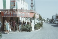 A group of people is gathered outside a restaurant with a large red awning that reads 'RESTAURANTE.' The building is located on a street lined with greenery and there are a few parked cars. The atmosphere appears relaxed, and the people are dressed in casual attire typical of a warm climate. A sign for 'Cruzcampo' beer is visible on the building.