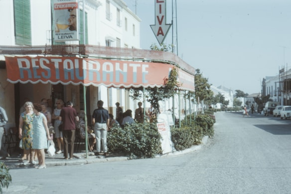 A group of people is gathered outside a restaurant with a large red awning that reads 'RESTAURANTE.' The building is located on a street lined with greenery and there are a few parked cars. The atmosphere appears relaxed, and the people are dressed in casual attire typical of a warm climate. A sign for 'Cruzcampo' beer is visible on the building.