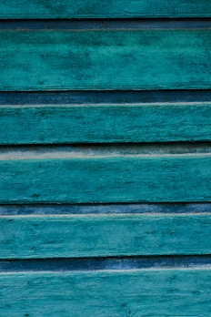 Weathered wooden panels painted in a vibrant teal color, displaying a rustic and textured surface with visible grain patterns and variations in the paint.