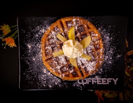 A round waffle sprinkled with powdered sugar, topped with sliced apples and a scoop of whipped cream, is placed on a black rectangular plate with the word 'COFFEEFY' written on it. Bright orange flowers with green stems are placed beside the plate.