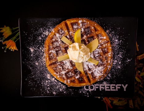 A round waffle sprinkled with powdered sugar, topped with sliced apples and a scoop of whipped cream, is placed on a black rectangular plate with the word 'COFFEEFY' written on it. Bright orange flowers with green stems are placed beside the plate.