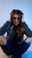 A person with long, wavy hair is sitting cross-legged, holding their head with both hands. They are wearing a denim jacket and ripped jeans, with a serene expression on their face.