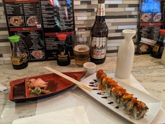 A sushi roll garnished with sauce and roe is elegantly presented on a white rectangular plate next to a small ceramic sake pitcher and cup. Nearby is a partially eaten meal with chopsticks resting on a red dish, accompanied by ginger and wasabi. A bottle and glass of Asahi beer are placed behind the meal, alongside soy sauce bottles. The scene is set on a marble counter with menu cards in the background.