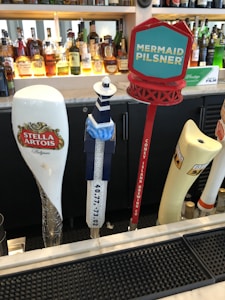 A bar counter features several beer tap handles, including brands such as Stella Artois, a lighthouse-shaped tap, and a tap labeled Mermaid Pilsner. In the background, a variety of liquor bottles are arranged on shelves.