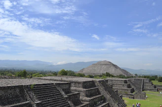 A large ancient pyramid structure stands prominently in a vast landscape. Foreground features stepped stone structures leading towards the pyramid. In the distance, mountains are visible under a sky with scattered clouds. Green vegetation surrounds the area, adding a natural contrast to the stone constructions. Small groups of people can be seen walking around the site, giving a sense of scale.