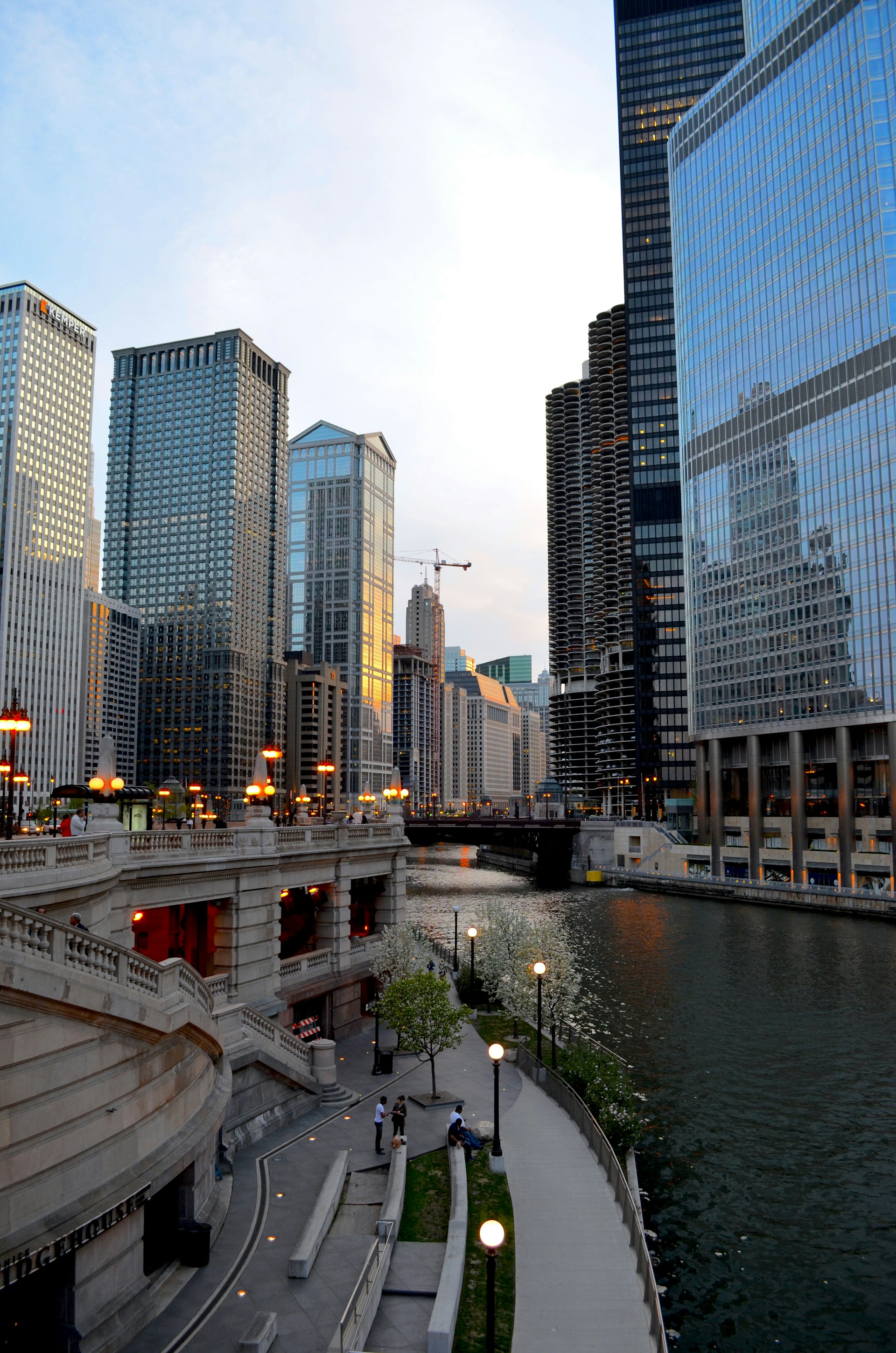 A scenic urban landscape featuring a river flanked by modern skyscrapers and an elegant stone walkway with ornate lampposts. The glass facades of the buildings reflect the soft evening light, and a few trees line the walkway. People are leisurely strolling along the path.