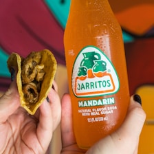 A person is holding a crispy taco filled with fried food in one hand and a glass bottle of Jarritos Mandarin soda in the other. The bottle is orange with condensation, indicating it is cold. The background features vibrant colors with a focus on a mural or painted wall.