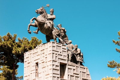 A detailed sculpture depicting a historical scene with figures on horseback atop a stone pedestal. The sculpture is intricately crafted with multiple figures, each in a dynamic pose. The scene is set against a clear blue sky, and trees are visible in the background, adding to the grandeur of the monument.