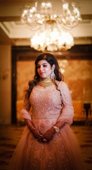 A woman dressed in an ornate, embellished gown stands elegantly beneath a chandelier. Her hands are adorned with intricate henna designs, and she wears statement jewelry, including large earrings and a necklace.