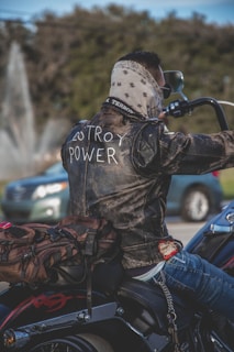 A person is riding a motorcycle wearing a leather jacket with the words 'Destroy Power' written on the back. The rider has a bandana covering their face and is carrying a brown backpack. In the background, there's a blurred car and trees, suggesting a street or park setting.