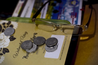 A pair of eyeglasses rests on a stack of papers, which include documents and a floral-patterned invitation. Several coins are scattered on top of the papers.