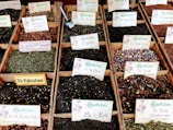 A variety of loose leaf teas displayed in wooden boxes, each with labels indicating different flavors and blends such as Té Verde Mediterraneo, Pu-Erh, and Té Pakistani. The teas have a range of colors and ingredients, with some featuring flower petals and spices. Metal scoops are visible in some of the boxes for serving.