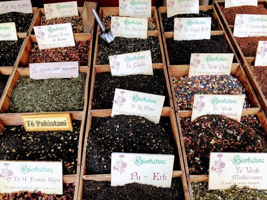 A variety of loose leaf teas displayed in wooden boxes, each with labels indicating different flavors and blends such as T&eacute; Verde Mediterraneo, Pu-Erh, and T&eacute; Pakistani. The teas have a range of colors and ingredients, with some featuring flower petals and spices. Metal scoops are visible in some of the boxes for serving.