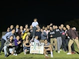 A large group of young adults is gathered on a grassy field at night. They are posing happily with various hand gestures, and one person is elevated on others' shoulders, holding a sign that reads 'Lambda Flex @ Winter Games.' Most of the people are casually dressed in sportswear.