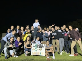 A large group of young adults is gathered on a grassy field at night. They are posing happily with various hand gestures, and one person is elevated on others' shoulders, holding a sign that reads 'Lambda Flex @ Winter Games.' Most of the people are casually dressed in sportswear.