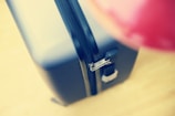 A close-up view of a hard-shell suitcase with a visible zipper and lock mechanism. The suitcase is positioned at an angle, emphasizing the smooth texture and sleek design. The background is blurred, and an indistinct red object is partially visible, adding contrast to the image.