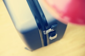 A close-up view of a hard-shell suitcase with a visible zipper and lock mechanism. The suitcase is positioned at an angle, emphasizing the smooth texture and sleek design. The background is blurred, and an indistinct red object is partially visible, adding contrast to the image.