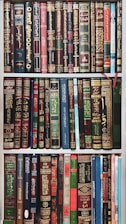A collection of books neatly arranged on a bookshelf. The books have ornate covers with elaborate designs and texts, predominantly in Arabic script. The books are of various colors and sizes, with some having bookmarks visible.