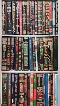 A collection of books neatly arranged on a bookshelf. The books have ornate covers with elaborate designs and texts, predominantly in Arabic script. The books are of various colors and sizes, with some having bookmarks visible.