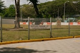 A parking lot area designated for suppliers is enclosed by a metal fence. A sign indicating 'SUPPLIERS PARKING LOT, 15 MINS GRACE PARKING ONLY' is prominently displayed on the fence. Beyond the fence, there are grassy areas with scattered large trees and some construction equipment, including barriers and a vehicle. The scene suggests a mix of parking and construction activities.