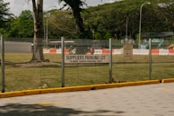 A parking lot area designated for suppliers is enclosed by a metal fence. A sign indicating 'SUPPLIERS PARKING LOT, 15 MINS GRACE PARKING ONLY' is prominently displayed on the fence. Beyond the fence, there are grassy areas with scattered large trees and some construction equipment, including barriers and a vehicle. The scene suggests a mix of parking and construction activities.
