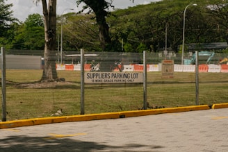 A parking lot area designated for suppliers is enclosed by a metal fence. A sign indicating 'SUPPLIERS PARKING LOT, 15 MINS GRACE PARKING ONLY' is prominently displayed on the fence. Beyond the fence, there are grassy areas with scattered large trees and some construction equipment, including barriers and a vehicle. The scene suggests a mix of parking and construction activities.