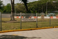 A parking lot area designated for suppliers is enclosed by a metal fence. A sign indicating 'SUPPLIERS PARKING LOT, 15 MINS GRACE PARKING ONLY' is prominently displayed on the fence. Beyond the fence, there are grassy areas with scattered large trees and some construction equipment, including barriers and a vehicle. The scene suggests a mix of parking and construction activities.