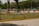 A parking lot area designated for suppliers is enclosed by a metal fence. A sign indicating 'SUPPLIERS PARKING LOT, 15 MINS GRACE PARKING ONLY' is prominently displayed on the fence. Beyond the fence, there are grassy areas with scattered large trees and some construction equipment, including barriers and a vehicle. The scene suggests a mix of parking and construction activities.