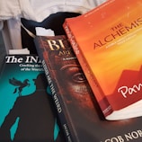 A close-up view of three books stacked together. The top book is 'The Alchemist' with an orange cover, featuring the name 'Paulo Coelho' prominently. The second book, underneath, has a dark, mysterious design with partial text visible. The bottom book titled 'The IN' has a silhouette of a man with a horse on the cover, set against a teal background.
