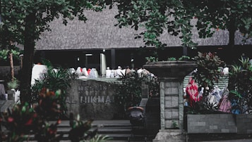 A group of people dressed in various colors of traditional attire gather outside a building labeled 'Masjid Al Hikmah'. The scene is framed by lush green trees and plants, with a stone pillar and other architectural elements visible in the foreground.