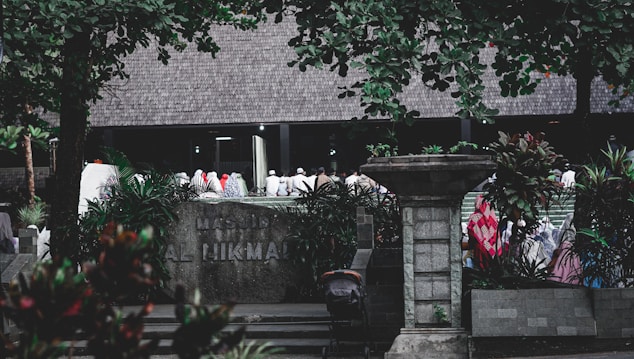 A group of people dressed in various colors of traditional attire gather outside a building labeled 'Masjid Al Hikmah'. The scene is framed by lush green trees and plants, with a stone pillar and other architectural elements visible in the foreground.