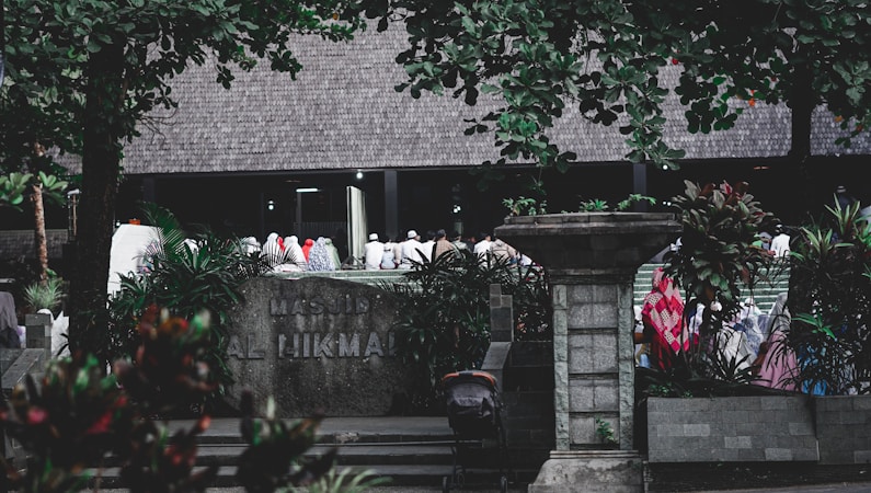 A group of people dressed in various colors of traditional attire gather outside a building labeled 'Masjid Al Hikmah'. The scene is framed by lush green trees and plants, with a stone pillar and other architectural elements visible in the foreground.