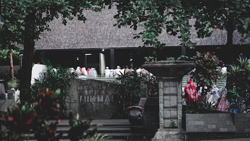 A group of people dressed in various colors of traditional attire gather outside a building labeled 'Masjid Al Hikmah'. The scene is framed by lush green trees and plants, with a stone pillar and other architectural elements visible in the foreground.