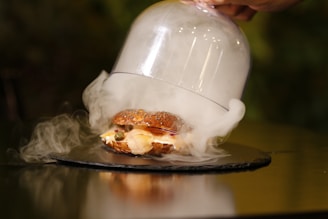A gourmet sandwich displayed under a glass cloche with mist or smoke swirling around it. The sandwich features various ingredients, including sliced meats and vegetables, on a seeded bun. The cloche is raised slightly, revealing the sandwich with an artistic presentation on a dark surface.