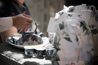 A hand is pouring tea from a blue and white patterned teapot into matching teacups. The scene is set on a wicker table, accompanied by floral-patterned cloth bags. Steam rises gently from the teapot, suggesting a freshly brewed tea session.