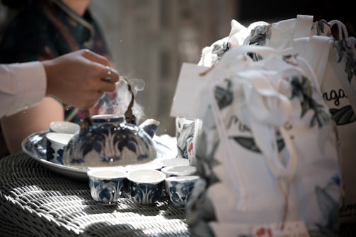 A hand is pouring tea from a blue and white patterned teapot into matching teacups. The scene is set on a wicker table, accompanied by floral-patterned cloth bags. Steam rises gently from the teapot, suggesting a freshly brewed tea session.