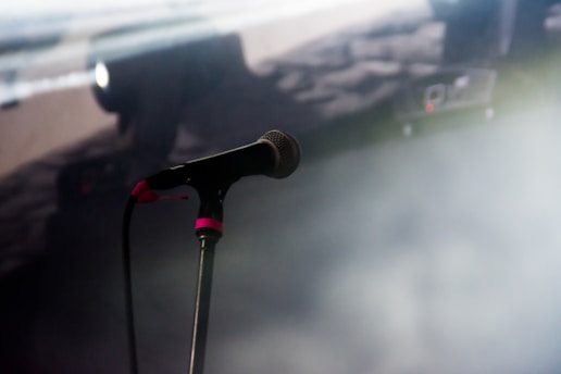 A microphone on a stand is positioned in a dimly lit environment. The background appears blurred with soft lighting, and there's a hint of fog or smoke, suggesting a stage setting.