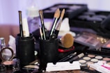 A variety of makeup tools and products are arranged on a surface. Brushes of different sizes are held in black cylindrical holders. A collection of compact powders, palettes, and other cosmetics are spread around. In the background, there is a faint outline of a water bottle.