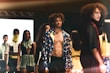 A fashion runway scene with models walking, showcasing diverse outfits in an indoor setting. The focus is on a shirtless male model wearing a colorful jacket, surrounded by other models displaying unique and stylish garments.